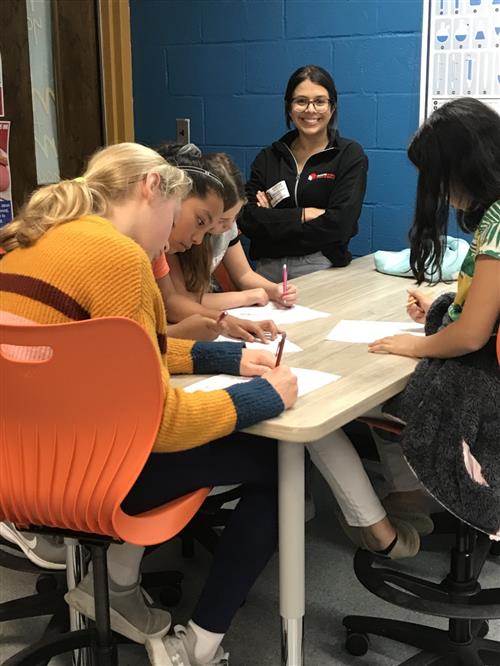 Woman mentor from University of MD looks on to students sitting at a table working on cyber security resource. 